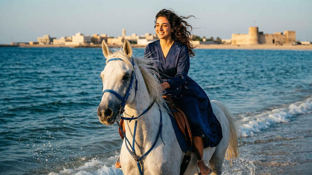 A person riding a white horse along a coastal shoreline with buildings in the background. 2