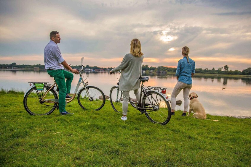 Three people on bicycles and a dog by a lake during sunset. 5