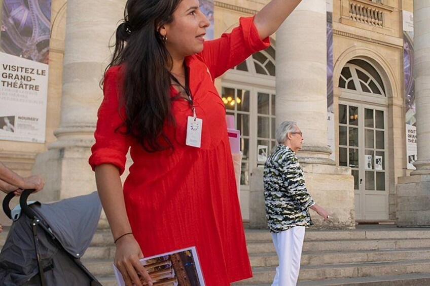 Una persona con un abito rosso tiene un libretto, in piedi di fronte a un edificio con colonne e un cartello con su scritto 'VISITEZ LE GRAND-THÉATRE'. 1