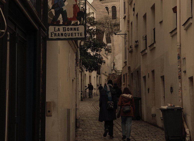 Eine enge Kopfsteinpflasterstraße mit Fußgängern, einem Schild mit der Aufschrift 'La Bonne Franquette' und einem Gebäude mit einer Kuppel im Hintergrund. 1