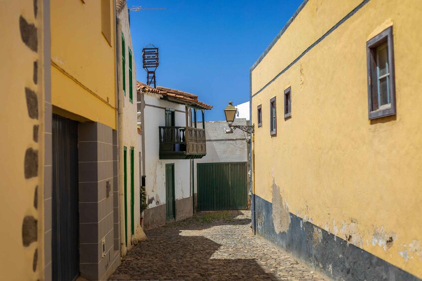 A narrow cobblestone alley flanked by buildings with yellow and white facades, a green door, and a small balcony. 2