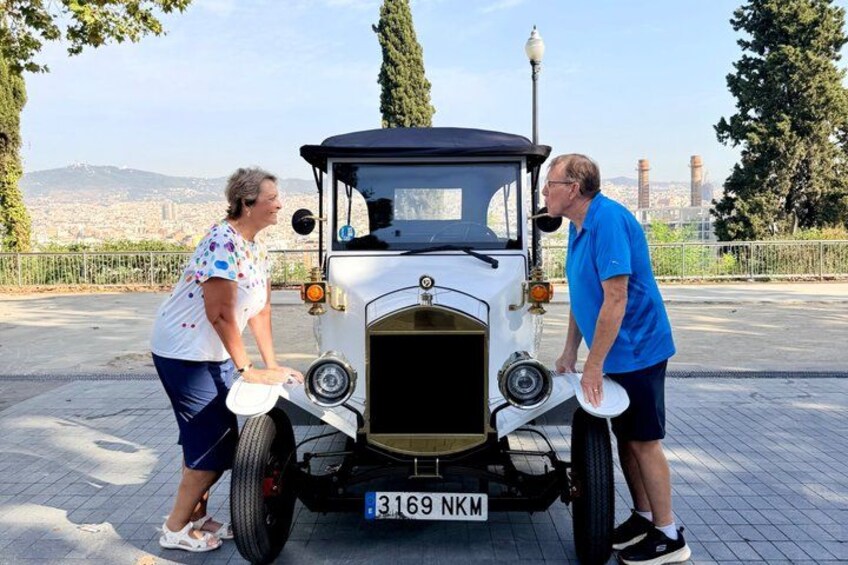 A vintage car with a license plate, two people interacting with it, and a cityscape in the background. 3