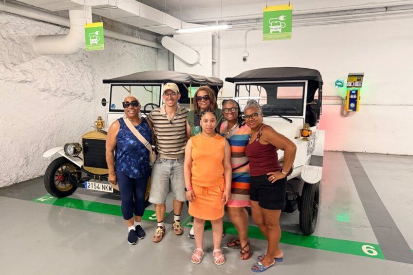 A group of people posing in front of a vintage car in a parking garage. 2