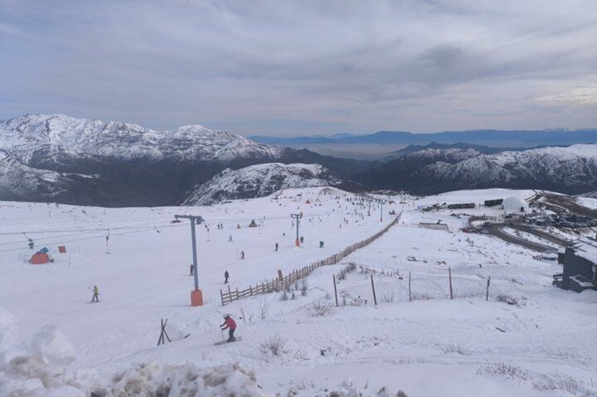 Pista de esquí nevada con esquiadores, un telesilla y una cordillera en el fondo. 2
