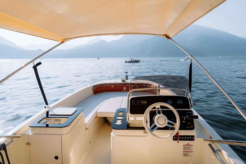 A boat's interior with a steering wheel, control panel, and a view of a calm lake with boats in the distance and mountains in the background. 3