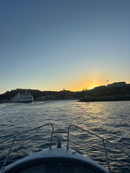 Veneen keulasta avautuu näkymä auringonlaskuun kaupungin yllä, ja horisontissa näkyy rakennuksia sekä suuri laiva. 1