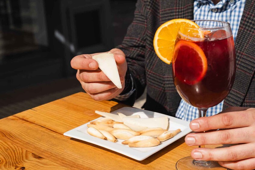 A person holding a glass of red drink with an orange slice, sitting at a wooden table with a plate of cheese slices. 3