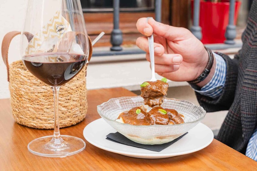 A person holding a spoon with food, a glass of red wine, and a woven basket on a table. 1