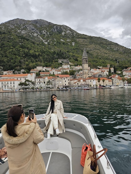 A person on a boat taking a photo with a smartphone, another person in a coat, and a coastal town with a prominent church in the background. 1
