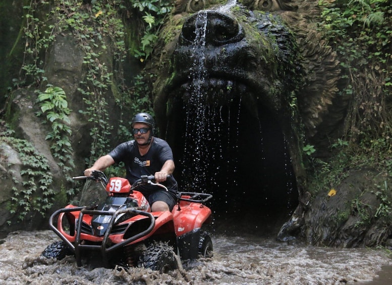 A person on an ATV navigating through a muddy stream with a waterfall in the background. 5