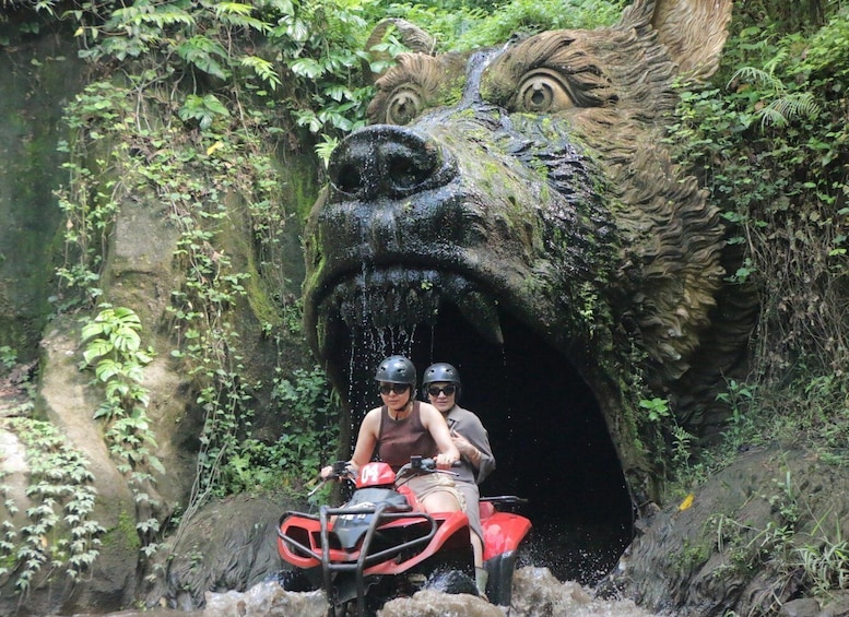Two people on a red ATV in a rocky tunnel with a large, moss-covered face sculpture. 1