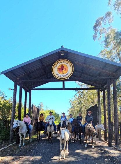 A group of people on horseback under a wooden shelter with a 'Maple Ranch Welcome' sign. 1