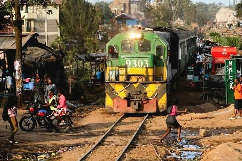 Un train portant le numéro 9303 sur sa façade, traversant une zone rurale où se trouvent des personnes et une moto à proximité. 1