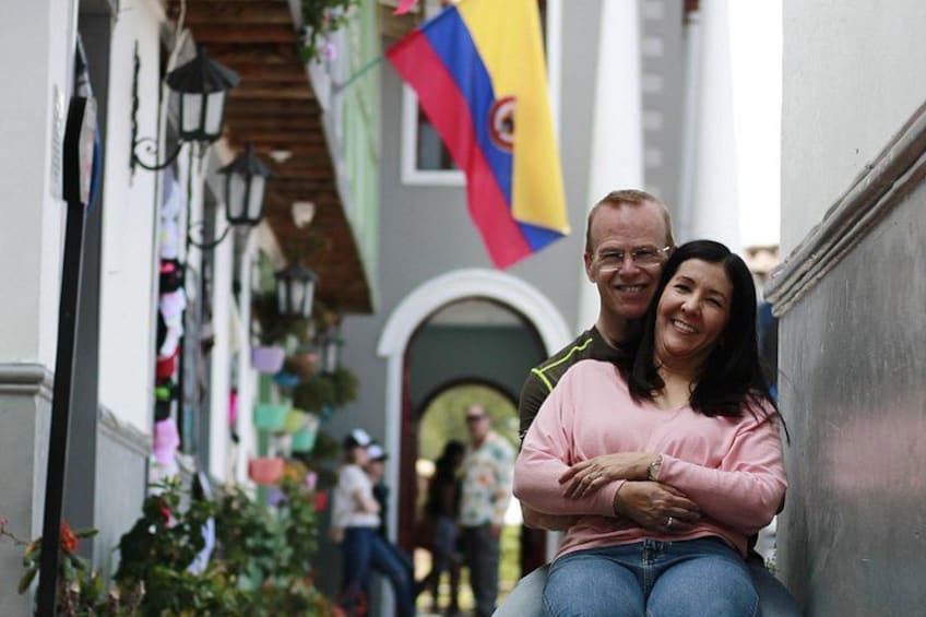A couple sitting on a bench in a narrow street with hanging plants and flags. 4