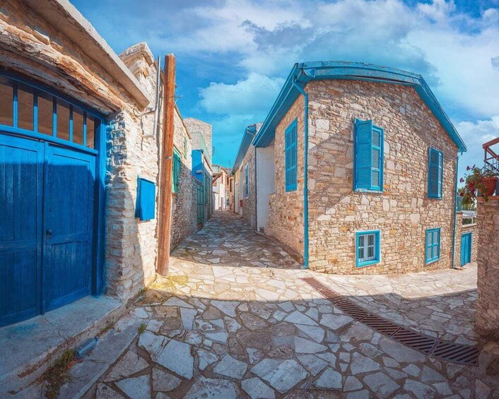 A narrow stone-paved street flanked by traditional stone buildings with blue shutters and doors. 1