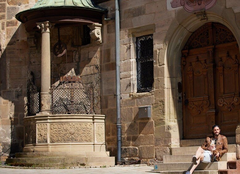 A stone building with a decorative fountain, a wooden door, and a person sitting on the steps. 2