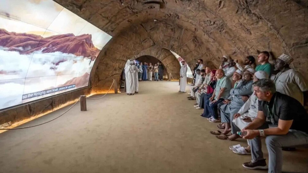 A group of people seated in a cave-like structure with a large mural depicting a mountainous landscape. 2