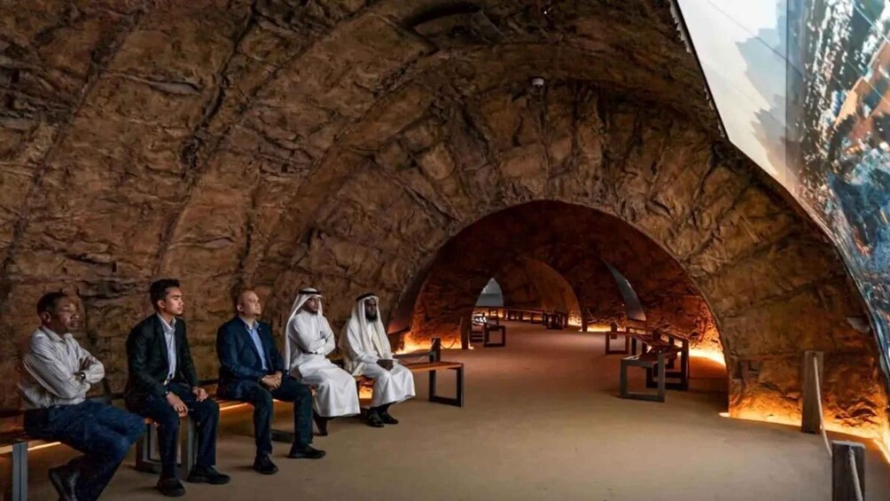 A group of people seated in a cave-like structure with a large mural on the right wall. 5