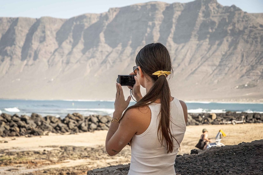 A person taking a photo with a camera, a rocky beach, and a mountain in the background. 1