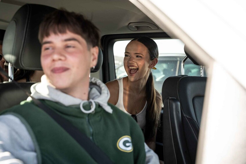 A person in a green sports jersey and another in a white tank top inside a vehicle. 2
