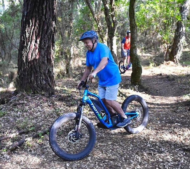 A person riding a blue bicycle with training wheels in a wooded area. 3