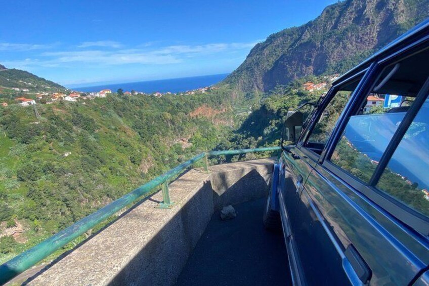 A car on a coastal road with a guardrail, overlooking a mountainous landscape and the sea. 2