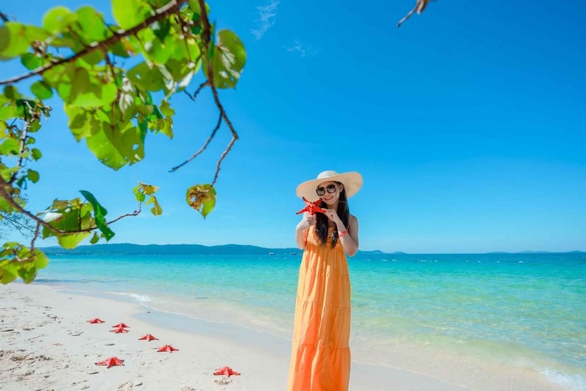 A person in an orange dress and white hat on a beach with clear blue water and distant mountains. 2