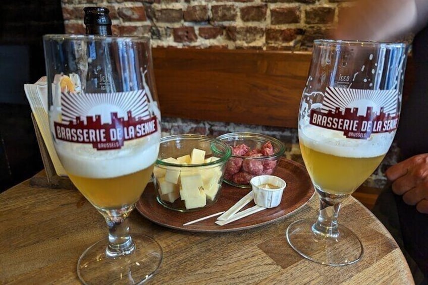 Twee bierglazen met 'Brasserie de La Senne' branding, een houten tafel en een schaal met kaas en fruit. 2