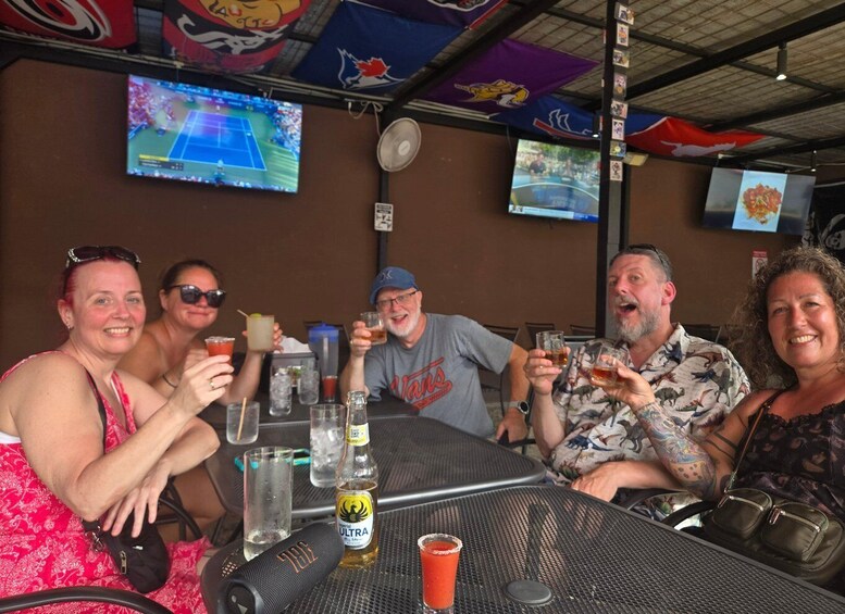 A group of people sitting at a table with drinks and a bottle of alcohol, in a room with sports team flags and a television screen. 3