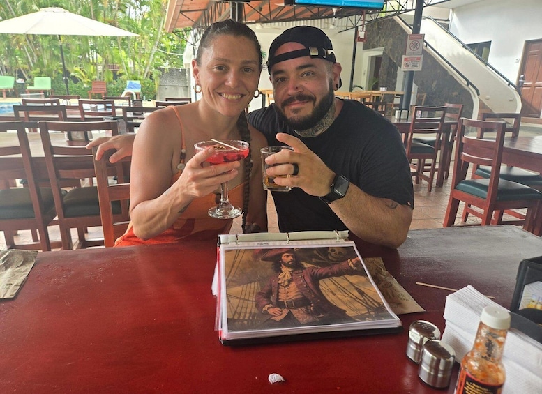 Two people sitting at a table with drinks, a book, and condiments. 2