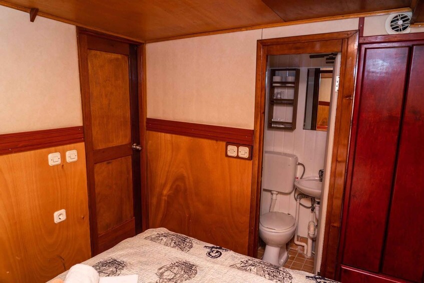 A small, compact room with wooden walls, a bed with patterned bedding, a toilet, and a cabinet. 4