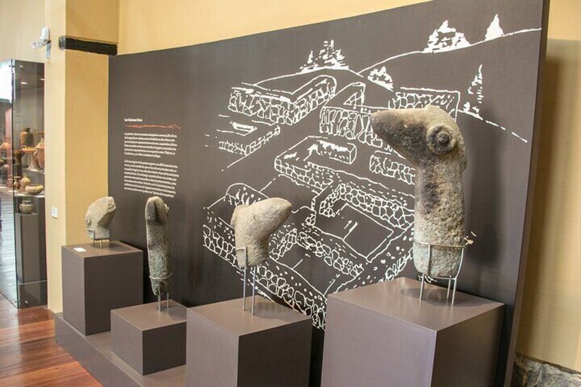 Une exposition dans un musée présentant d’anciens objets en pierre, disposés sur des piédestaux, et un mur orné d’un dessin au trait blanc représentant un établissement. 2