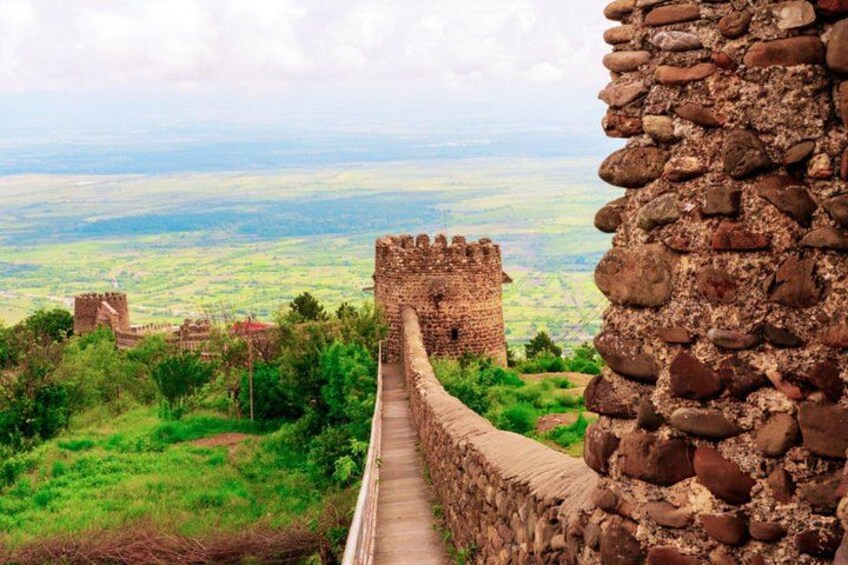 A stone wall with a pathway leading to a distant castle, surrounded by greenery and a hilly landscape. 3