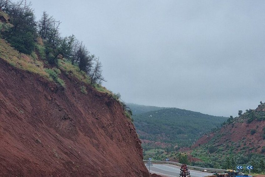 A steep red hillside with sparse vegetation, a winding road, and distant green hills. 5