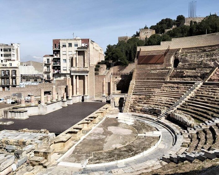 An ancient amphitheater with stone seating and a central stage, surrounded by modern buildings and a hill with ruins. 5