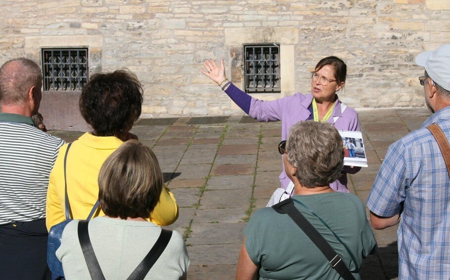 A person gesturing while speaking to a group of people in front of a stone building with barred windows. 2