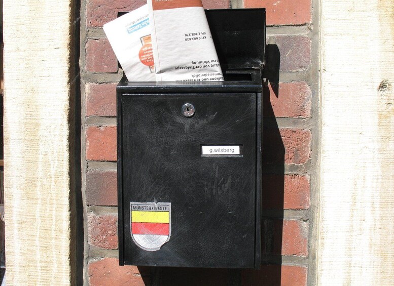 A black mailbox with a sticker of Münster/Weste and a nameplate reading 'g.wilsberg' against a brick wall. 5