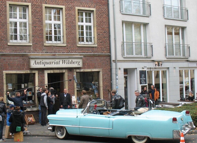 A light blue vintage car parked in front of a building with a sign reading 'Antiquariat Wilsberg.' 3