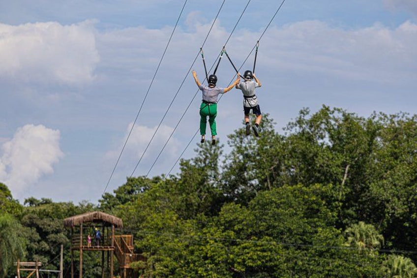 Two people ziplining over a forested area with a wooden platform in the background. 5