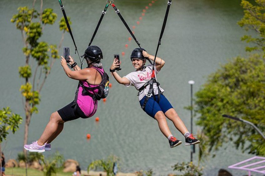 Two people on a zip line, holding onto handles, with a body of water and trees in the background. 4