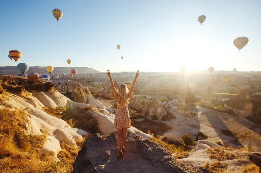 A person standing on a rocky outcrop with arms raised, overlooking a landscape with numerous hot air balloons in the distance. 4