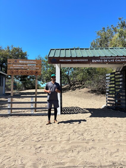 A man standing in front of a wooden sign with 'Bienvenidos Monumento Natural Las Dunas de las Calderas' and 'Welcome / Bienvenidos' written on it. 2
