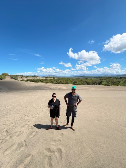 Two people standing on a sandy beach with footprints, a clear sky, and distant hills. 5