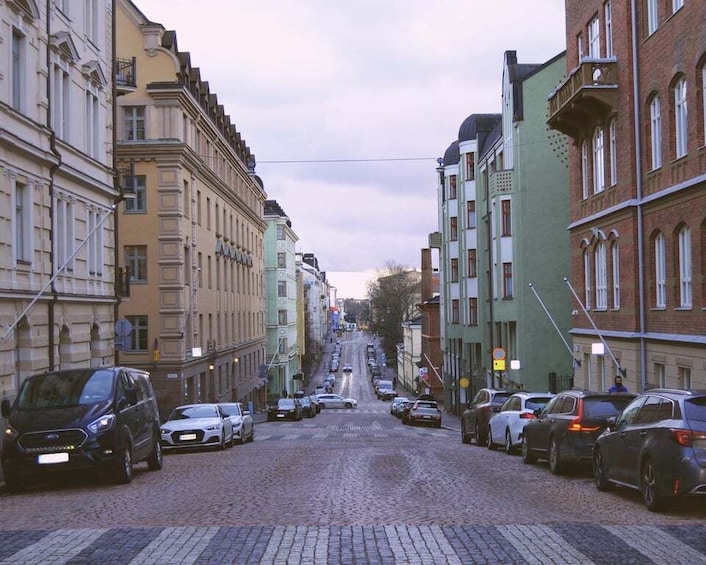 A cobblestone street lined with multi-story buildings, parked cars, and a clear sky. 4
