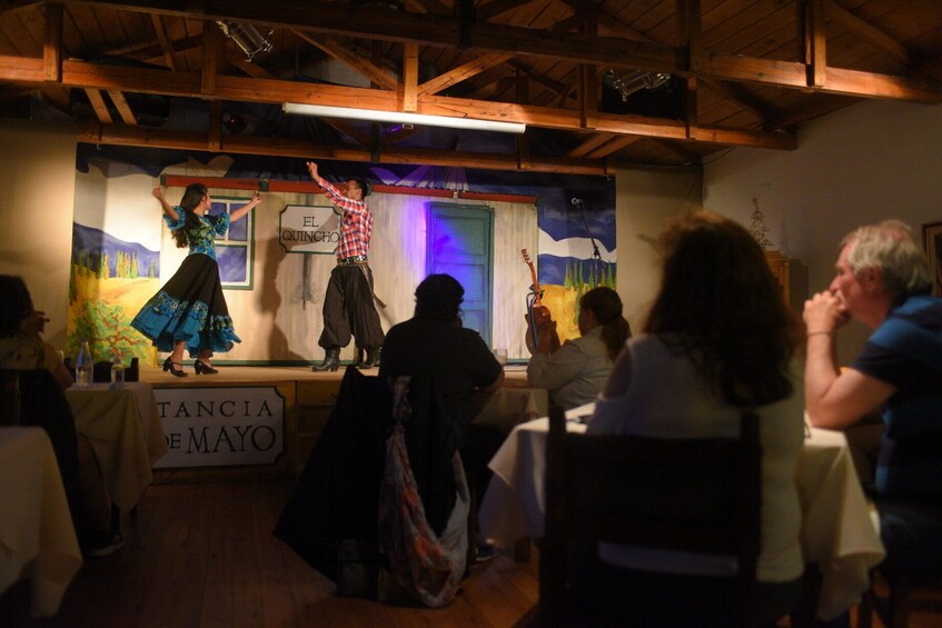 A stage with performers in traditional attire, a sign reading 'EL QUINCHO,' and a banner for 'TANCIA DE MAYO.' 1