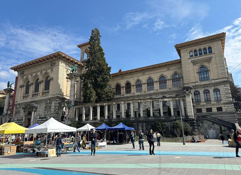 A historic building with classical architecture, a central tree, and outdoor market stalls. 2