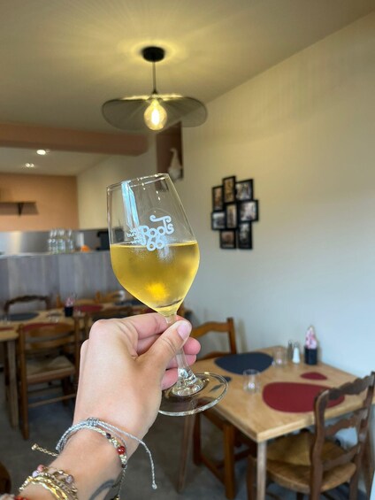 A hand holding a glass of white wine with 'bunk the Roots' etched on it, in a dining area with wooden tables and chairs. 4