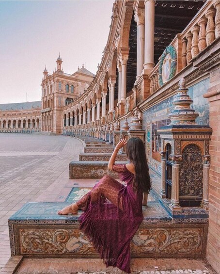 A woman in a purple dress sitting on steps with ornate architectural details and a large building in the background. 2