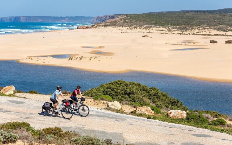 Two cyclists on a coastal road with a beach and cliffs in the background. 1