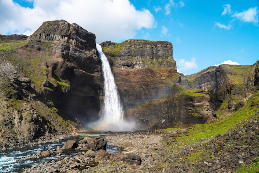 Een waterval die naar beneden stort over een rotswand, omringd door weelderige begroeiing en een helderblauwe lucht. 1